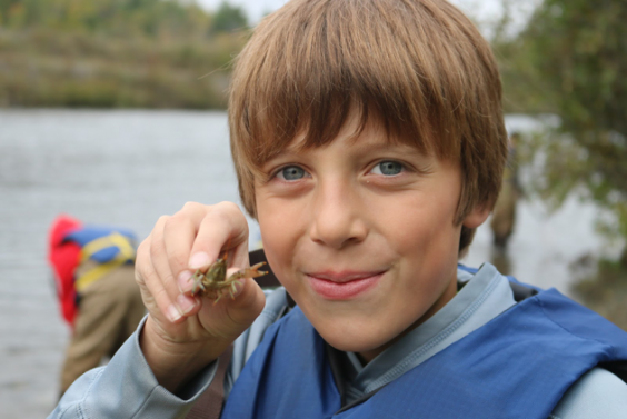 A NEMIGLSI student holds a crayfish up to the camera.  Behind the student a lake and trees can be seen out of focus, along with one person looking into the water.