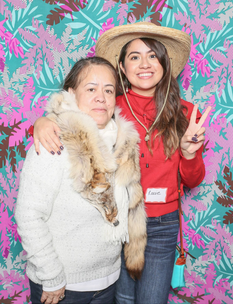 Rosalba Herrera posing against a vibrant background, one hand giving a peace sign, the other draped over the shoulders of an unidentified older woman.