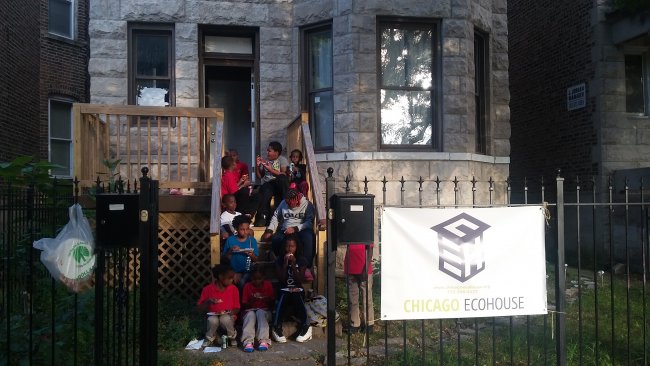 Participating youth sitting on the steps of Chicago Eco House. A sign on a wrought iron gage reads Chicago Eco House. Through the open gate entrance 10 children can be seen sitting on wooden steps up to a wooden deck off the the stone house, and beside its protruding bay window. The children are eating pizza.